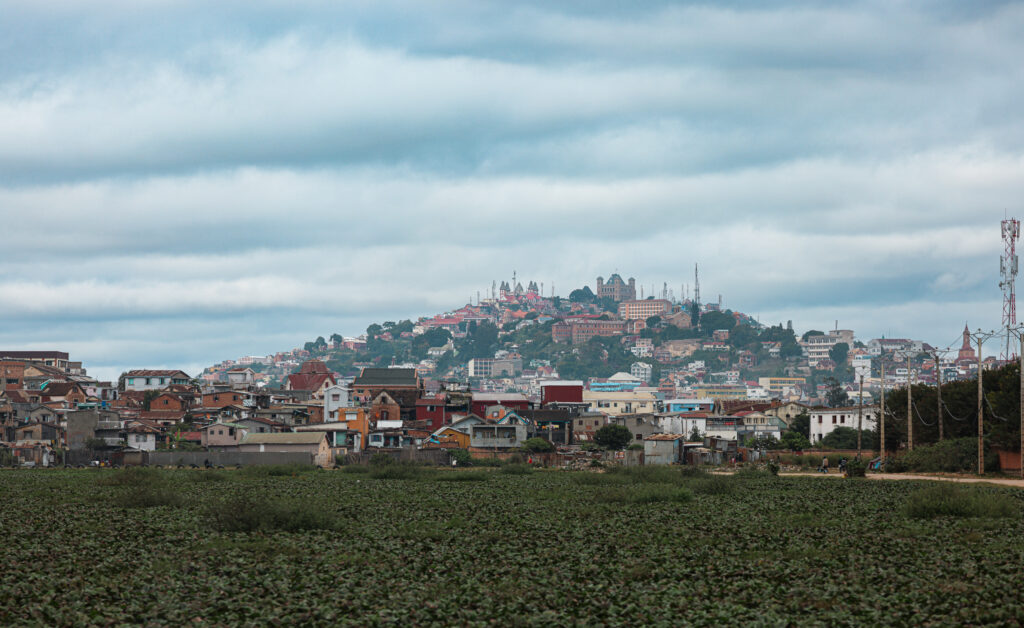 A vibrant suburb of Antananarivo showcases houses in various colors, set against a backdrop of rolling green fields and an overcast sky, creating a picturesque urban landscape.